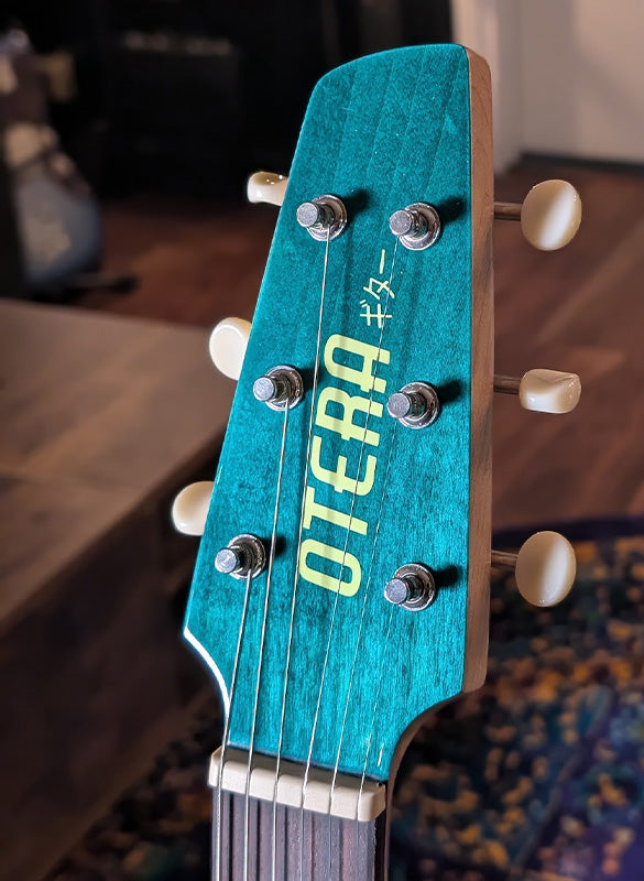 Close-up of a teal guitar headstock with 'Otera' branding on a blurred background.